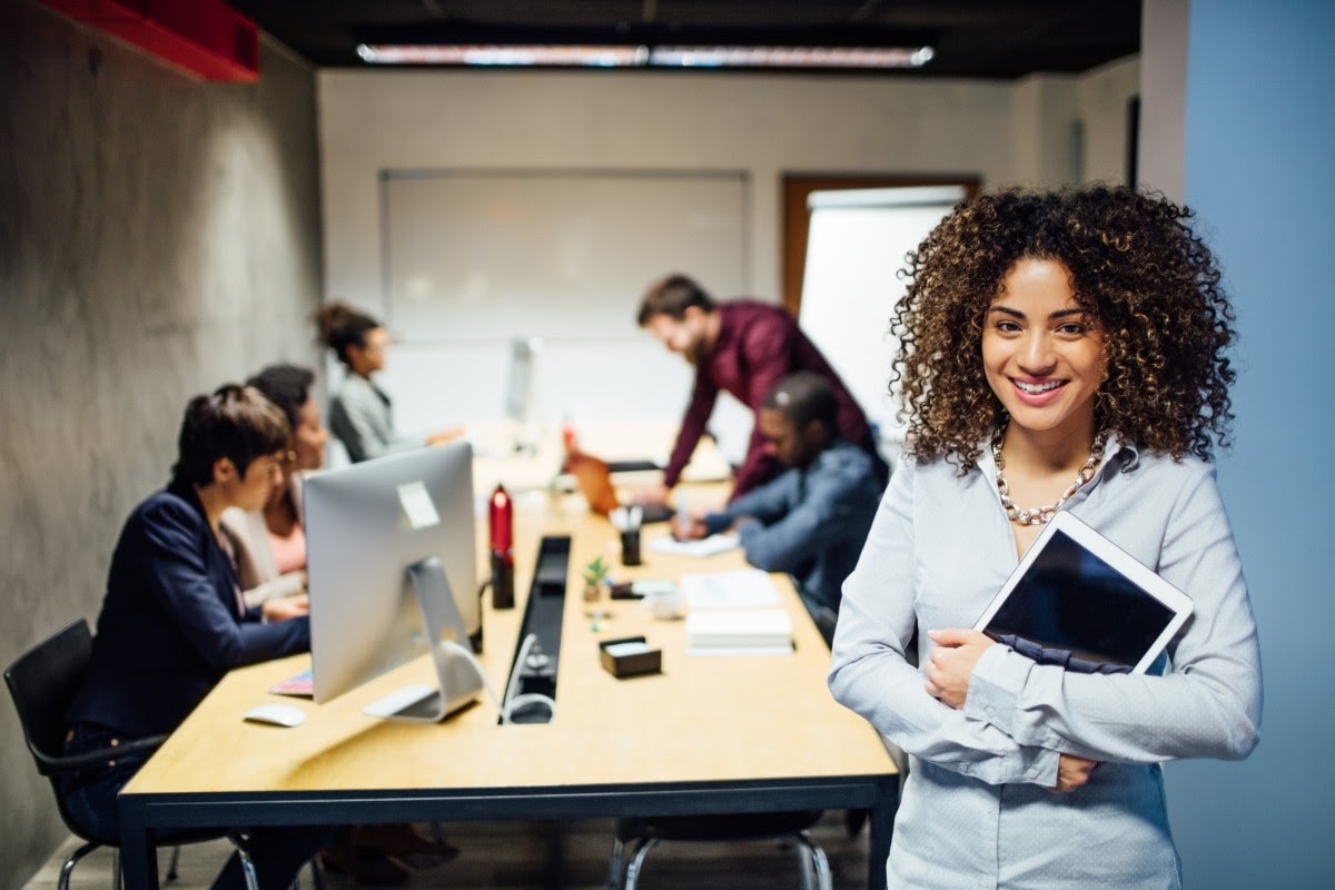 Estagiária feliz segurando tablet, com equipe ao fundo